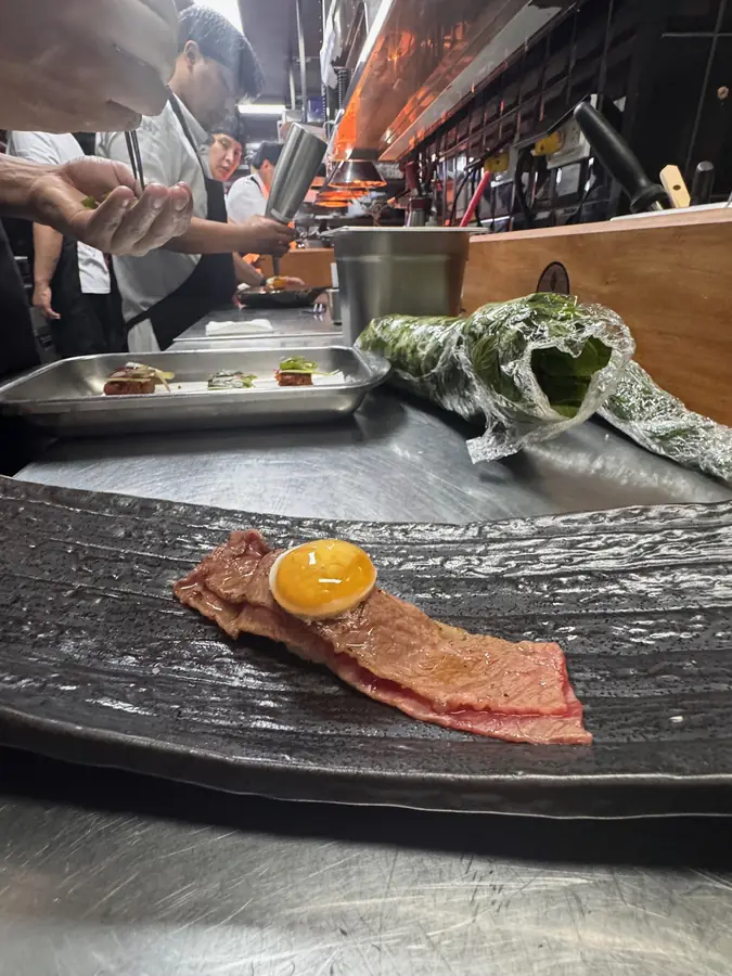 Thin slices of beef topped with an egg yolk on a dark plate, cooks plating behind