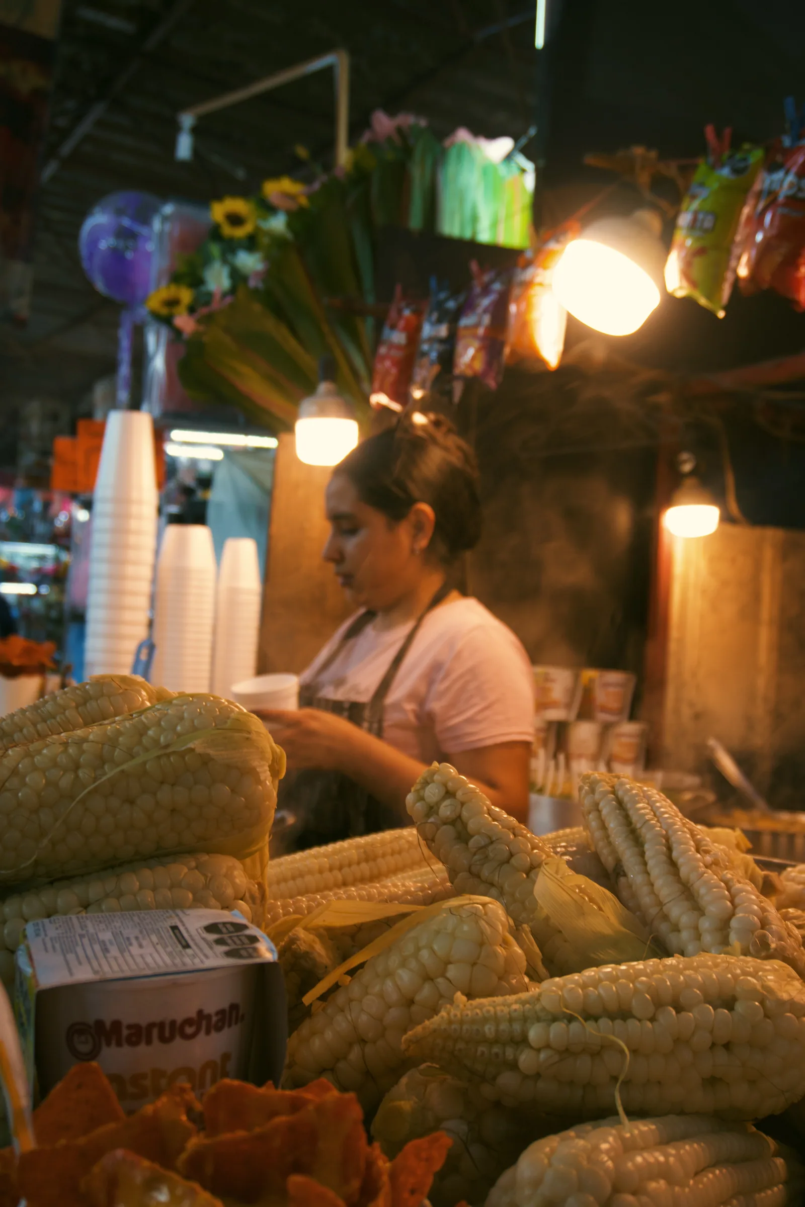A vendor behind ears of corn and containers of ingredients at a market stall